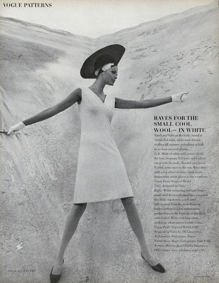 Black and white photo of a model in a v-neck bias dress worn with hat, gloves, and patent-leather shoes against a barren landscape
