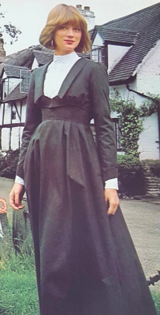 1970s Laura Ashley catalogue photo of a model in a long-sleeved jacket, white blouse, and matching ankle-length black jumper, seen outdoors in front of a Tudor-style house