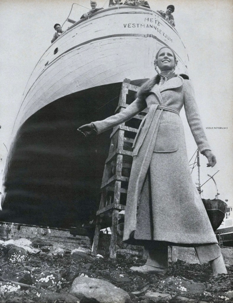 A model in a knit wrap coat stands on a rocky Icelandic beach before the prow of a ship and a rustic ladder, with young people looking down from the ship