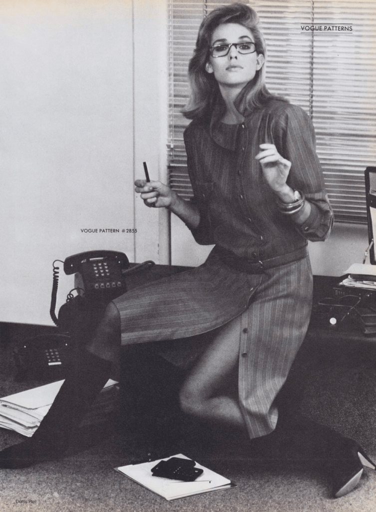 A model, wearing glasses, lookes up, wearing a dress made using Vogue 2855 by Chloe, seated on a low bench between a black telephone and an open briefcase. Another telephone is on the floor beside a stack of legal-size file folders, and a calculator sits on the floor in front of her.